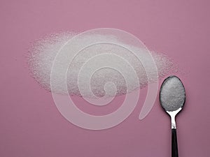 Heap of white sugar with a spoon filled with sugar on a pink background top view