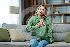 Heart attack, panic attack,. An elderly woman sits on the sofa at home and holds her hand to her chest. It is difficult