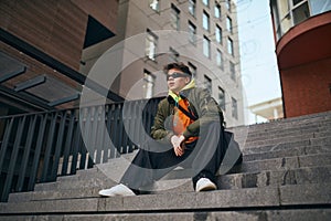 In heart of city& x27;s rhythm. Cool attitude young man sitting with thoughtful expression on stairs against skyline