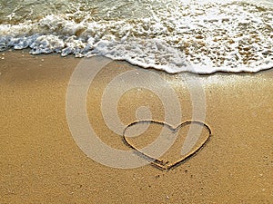 Heart drawn on the sand of a sea beach with white wave at sunset