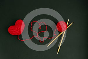 The heart is knitted of red threads on a dark background with a ball and wooden knitting needles.