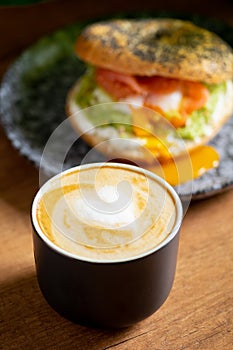 Heart latte art coffee scene with a bagel with egg, avocado, and fish on a wooden table. Perfect breakfast or brunch