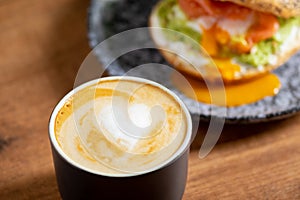 Heart latte art coffee scene with a bagel with egg, avocado, and fish on a wooden table. Perfect breakfast or brunch