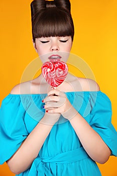 Heart Lollypop. Beautiful teen girl with colourful lolli pop. Funny teenage with bow hairstyle isolated on studio yellow