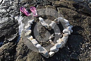 Heart made of stone on the beach with pink and violet flip flops next to it