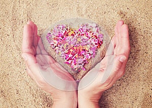 Heart of pink petals of tropical flowers on the sand and female hands, close-up