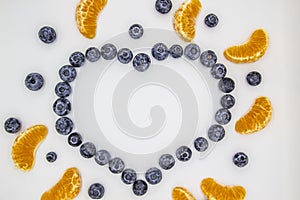A heart`s frame from mandarin pieces and blueberry on white background. A composition of the mandarin pieces and berries in form