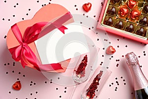 Heart-shaped box with blank paper card, box of chocolates, bottle of champagne and glasses with confetti on pink background.