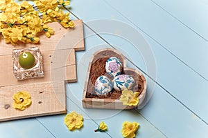 Heart shaped box with decorated Easter eggs, yellow flowers, burning candle and brown boards on wooden blue background. Spring
