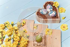 Heart shaped box with decorated Easter eggs, yellow flowers, burning candle and brown boards on wooden blue background. Spring