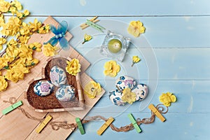Heart shaped box with decorated Easter eggs, yellow flowers, burning candle and brown boards on wooden blue background. Spring and