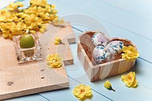 Heart shaped box with decorated Easter eggs, yellow flowers, burning candle and brown boards on wooden blue background. Spring