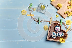 Heart shaped box with decorated Easter eggs, yellow flowers, burning candle and brown boards on wooden blue background. Spring