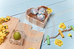 Heart shaped box with decorated Easter eggs, yellow flowers, burning candle on wooden blue background. Spring and Easter