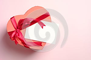 Heart shaped box with red ribbon bow and blank paper card on pink background. Flat lay, top view. Valentine`s day, Mother`s day,