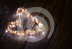 A heart shaped figure laid out light LED garland on vintage brown wooden board. Heart made of LED rice lights. Concept of