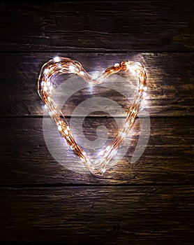 A heart shaped figure laid out light LED garland on vintage brown wooden board. Heart made of LED rice lights. Concept of