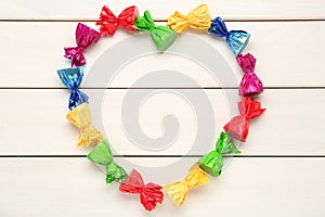 Heart shaped frame of candies in colorful wrappers on beige wooden table, flat lay. Space for text