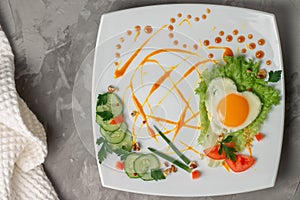 Heart shaped fried egg with vegetables, herbs and sauce on square white plate on gray concrete background. Flat lay