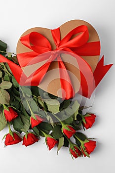 Heart shaped gift box with bow and beautiful red roses on white background, flat lay