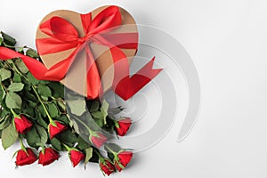 Heart shaped gift box with bow and beautiful red roses on white background, flat lay. Space for text