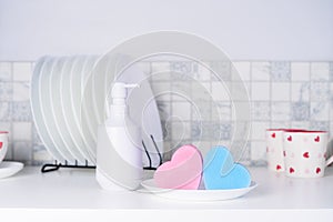 Heart shaped sponges, dish wash soap in the white container and clean plates and cups on the kitchen table. Modern