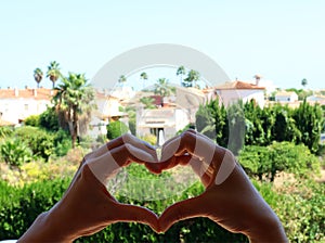 Heart symbol from hands on background of Spanish landscape, greenery, palm trees, houses, love for Spain