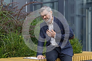 Heartache of mature man, gray haired businessman holding hands on rough, heart attack pain, boss outside modern office