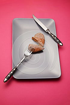 Heartbreaker concept image: heart shaped cookie on plate with fork and knife over pink background photo