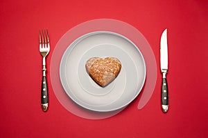 Heartbreaker concept image: heart shaped cookie on plate with fork and knife over red background photo