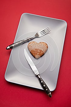Heartbreaker concept image: heart shaped cookie on plate with fork and knife over red background photo