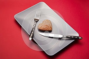 Heartbreaker concept image: heart-shaped cookie on plate with fork and knife over red background photo