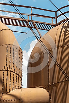 Heavily riveted metal tanks and catwalk, industrial shapes against a blue sky with clouds