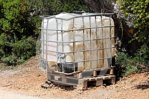 Heavily used old white intermediate bulk container or IBC plastic tank with twisted cracked metal cage on top of wooden pallets