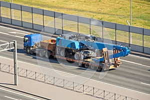 Heavy blue excavator long boom bucket on transportation truck with rubber wheels long trailer platform on the highway in the city