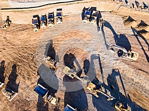 Heavy dump trucks, bulldozers, and excavators on yellow clay construction site. Long shadows on the ground. Top view at sunset