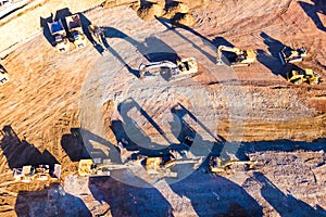 Heavy dump trucks, bulldozers, and excavators on yellow clay construction site. Long shadows on the ground. Top view at sunset