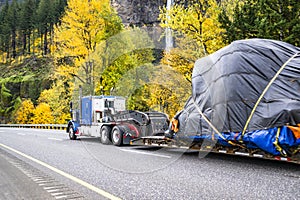 Heavy duty blue big rig classic American semi truck transporting covered oversized cargo driving on the autumn road with yellow