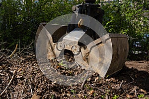 Heavy industrial machine scoop wooden branches and dirt of the ground. Hydraulic shovel lifting wood and waste from the garden