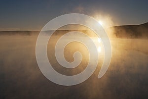 A heavy mist hangs over a calm lake at sunrise