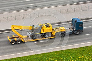 Heavy new yellow excavator long boom bucket on transportation truck with rubber wheels long trailer platform on the highway in the