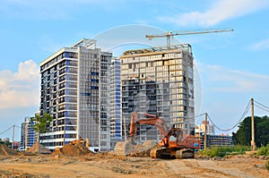 Heavy tracked excavator at a construction site on a background of a residential building and construction cranes on a sunny day