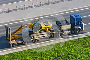 Heavy yellow excavator on transportation truck with long trailer platform on the highway in the city