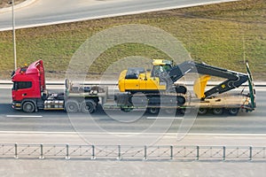 Heavy yellow excavator on transportation truck with long trailer platform on the highway in the city
