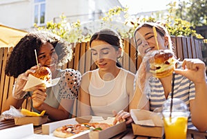 heerful happy female friends of different nationalities snacking on burgers and pizza outdoors and having fun together