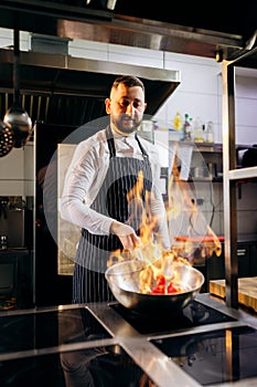 Ð¡hef in the professional kitchen with a frying pan and a fire..Chef`s hands hold iron Pan and preparing food on cooker