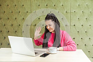 Hello. Affable young happy woman with black dreadlocks hairstyle in pink blouse sitting and greeting worker through a webcam at