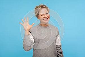 Hello! Cheerful woman with short hair in casual sweatshirt smiling friendly at camera and waving hi, welcoming