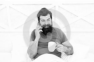 Hello dear. Bearded man using mobile technology in bed. Handsome guy talking on phone and drinking coffee at home