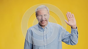 Hello. Happy excited sociable senior man waving hand to camera and smiling, greeting cheerfully, yellow background